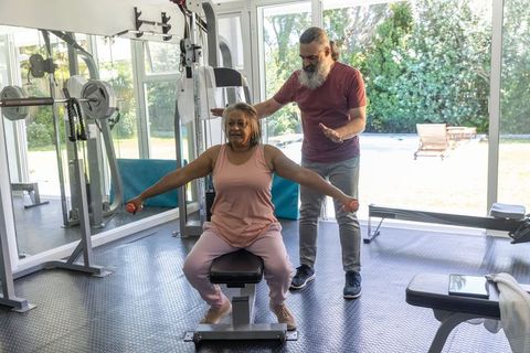 Senior african american woman doing seated lateral raises with trainer in home gym