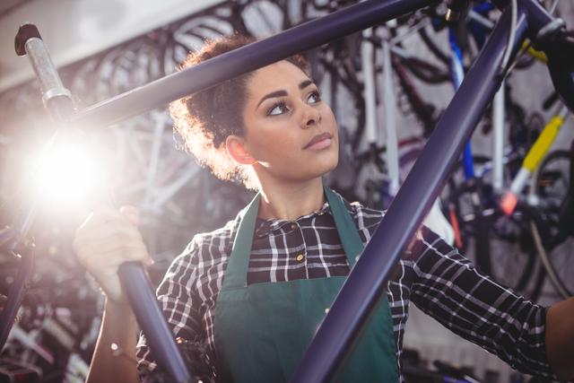 Female mechanic holding a bicycle frame in a workshop filled with various bikes. She is focused on her work, wearing a plaid shirt and an apron. Ideal for use in articles or advertisements related to bicycle repair services, women in trades, or cycling maintenance tutorials.