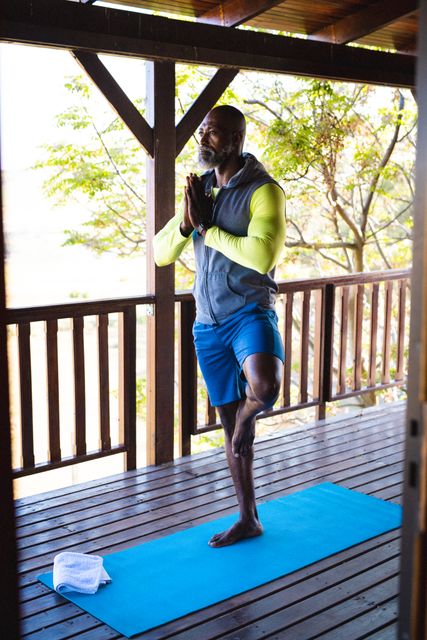Senior African American man practicing tree pose on exercise mat on balcony at log cabin. Ideal for content related to fitness, active lifestyle, yoga, meditation, retirement, wellness, and vacation. Perfect for promoting healthy living and solitude in nature.