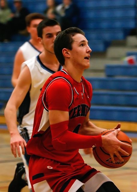 Young male basketball player in red jersey focused on scoring while holding the ball. Teammate and opponent visible in background, with blue bleachers suggesting a gymnasium setting. Useful for sports-related content, teamwork, athleticism, or goal-oriented themes.