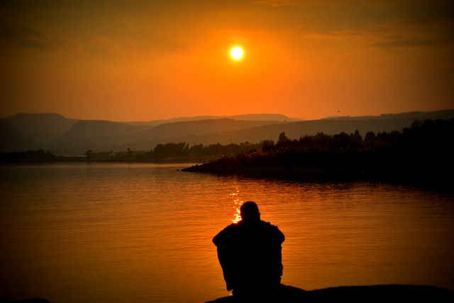 Person sitting alone by lake watching vibrant sunset. Silhouette creates calming atmosphere perfect for themes of solitude, peace, and mindfulness. Useful for meditation, relaxation concepts, travel publications, and nature-related projects.
