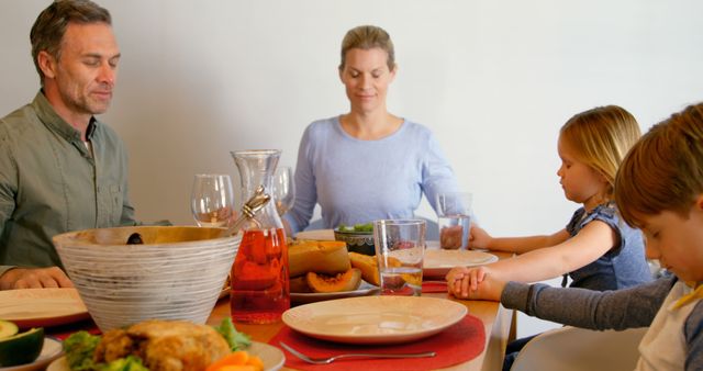 Family sitting together at dining table with eyes closed, holding hands in prayer before meal. Represents tradition, togetherness, and gratitude. Perfect for articles on family values, dining traditions, and religious observances.