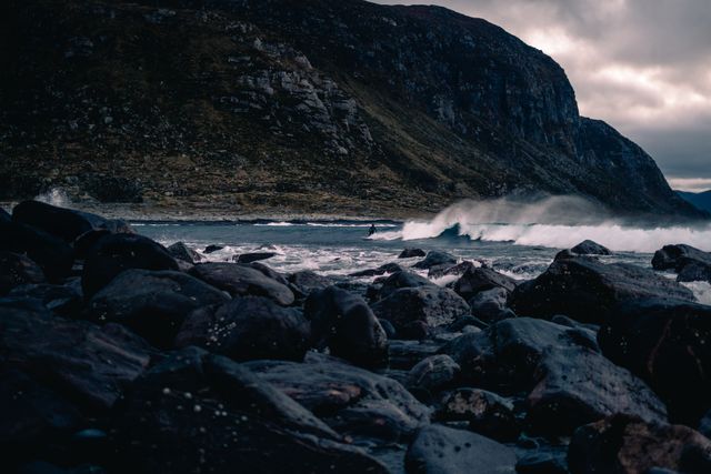 Photo showcases a rugged coastal landscape with dark, dramatic rocks and crashing waves under a moody sky. Suitable for themes relating to nature photography, wilderness, and coastal erosion. Great for use in travel blogs, outdoor adventure websites, and environmental conservation promotions.