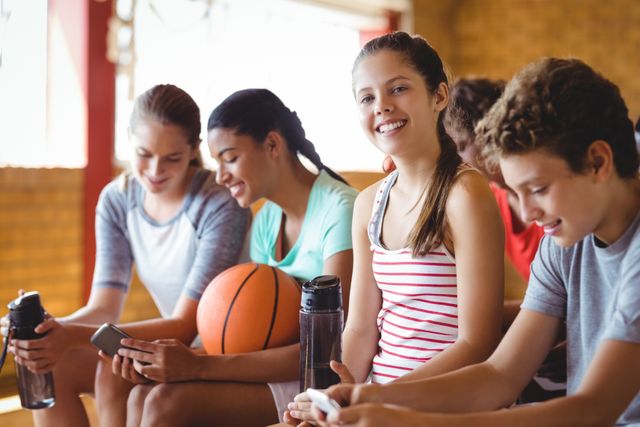 Smiling high school kids using mobile phone while relaxing in basketball court