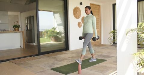 Asian woman performing weighted lunge on sunlit patio with dumbbells and yoga mat