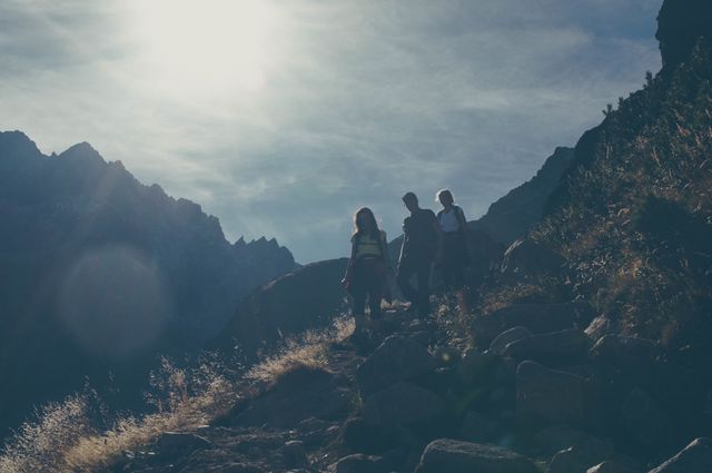 Group of hikers are trekking along a rocky mountain trail during daylight. The background features mountains and a cloudy sky, creating a sense of adventure and exploration. This image is suitable for use in travel brochures, outdoor activity promotions, fitness advertisements, and adventure blogs.