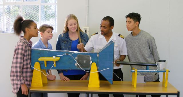 A diverse group of teenagers is engaged in a learning activity with a pilot, who is explaining the mechanics of an airplane model. Their attention and curiosity are evident as they listen and observe the demonstration, highlighting the importance of hands-on learning in education.