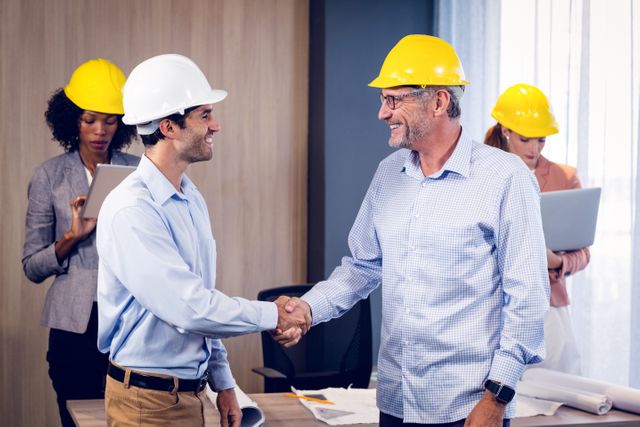 Architects shaking hands after a successful meeting in an office with colleagues working in the background. Ideal for illustrating concepts of teamwork, collaboration, successful business agreements, and professional partnerships in the construction and engineering industries.