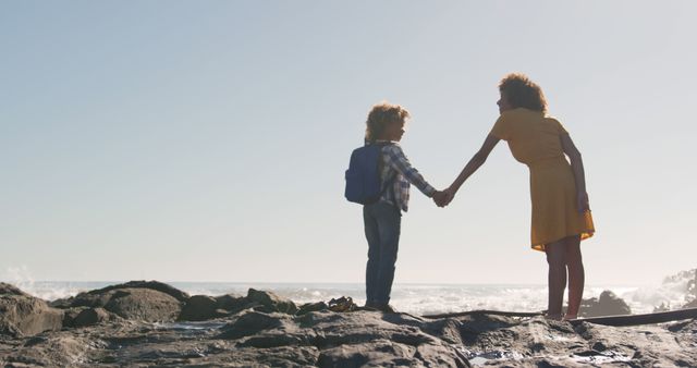 Happy biracial mother and son holding hands standing on sunny rocky beach, copy space. Motherhood, childhood, togetherness, summer, vacations and free time, unaltered.