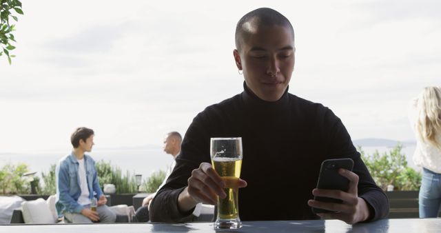 Young Asian woman sitting at an outdoor bar, holding a glass of beer, and using her smartphone. People in the background create a social and relaxed atmosphere. Ideal for use in lifestyle, technology, and social interaction contexts, as well as promoting outdoor dining and leisure activities.