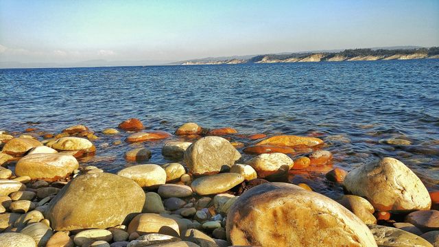 Rocky shoreline with smooth pebbles meeting clear blue sea under bright sky. Ideal for use in projects related to travel, nature, coastal appreciation, summer and tranquility. Perfect for postcards, vacation brochures, websites about nature destinations, or relaxing wallpapers.