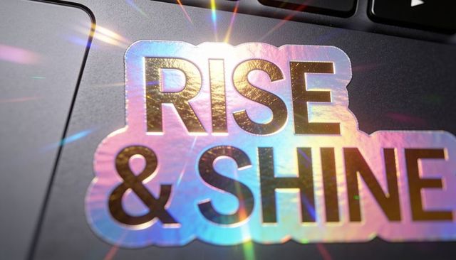 Holographic rise & shine sticker reflecting rainbow flares on laptop palm rest