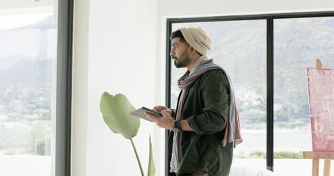 Indian designer sketching on tablet by window in bright minimalist studio with easel