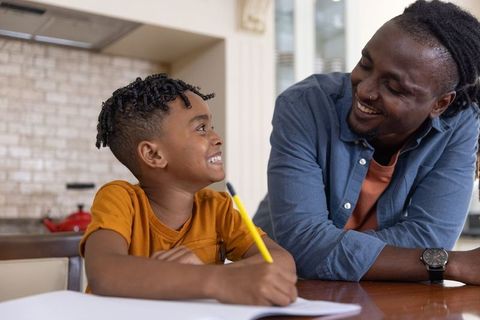 Smiling african american dad helping son with homework in kitchen, family bonding moment
