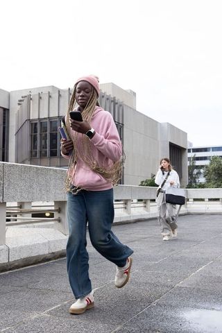 Young student walking on campus bridge checking phone carrying books in pink hoodie