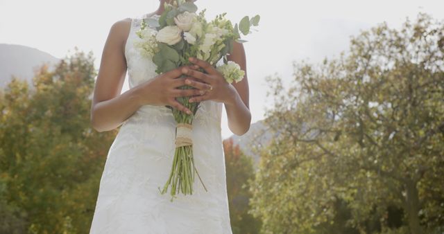 Bride shown holding a bouquet of white flowers, wearing a lace wedding dress, in an outdoor setting surrounded by trees and natural light. Ideal for wedding invitations, bridal magazines, and event planning materials.