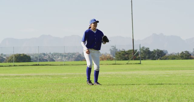 Youth baseball player in full uniform practicing on a grassy field. Mountains are visible in the background under a clear sky, creating a picturesque setting. Ideal for uses related to sports, youth activities, baseball training programs, athletic promotion, or outdoor recreation.