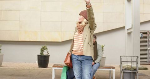 Mature woman hailing ride while holding shopping bags and brown satchel in city square