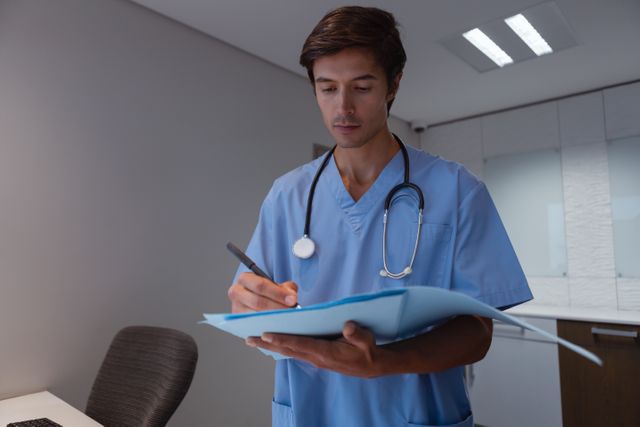 Male surgeon in blue scrubs writing on a medical report in a hospital office. Ideal for use in healthcare, medical, and hospital-related content, showcasing professional medical documentation and patient care.