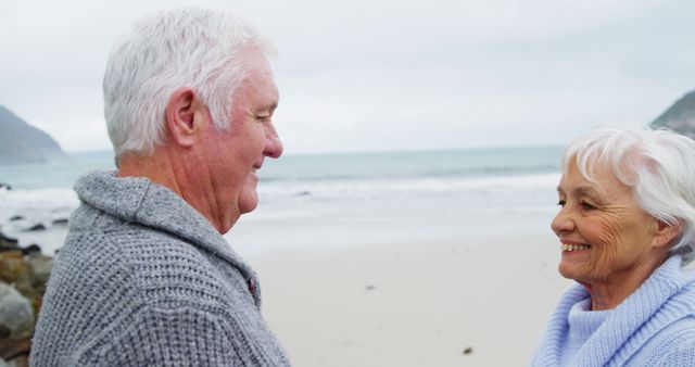 Elderly couple enjoying a moment at the beach, smiling warmly at each other. The ocean and mountains provide a serene backdrop, making this ideal for themes of aging, romance, retirement, and well-being. Perfect for advertisements, blogs, and articles focusing on happy relationships, outdoor activities, healthy aging, and beach vacations for seniors.