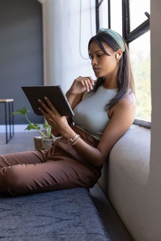 Asian woman using tablet and earbuds by window in modern cozy minimal workspace