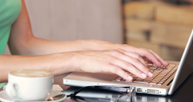 Image shows a woman’s hands typing on a laptop keyboard in a café, with a cup of coffee and a pair of eyeglasses beside them. Could be used for illustrating topics related to freelancing, remote work, business, technology, online communication, productivity, and casual working environments.