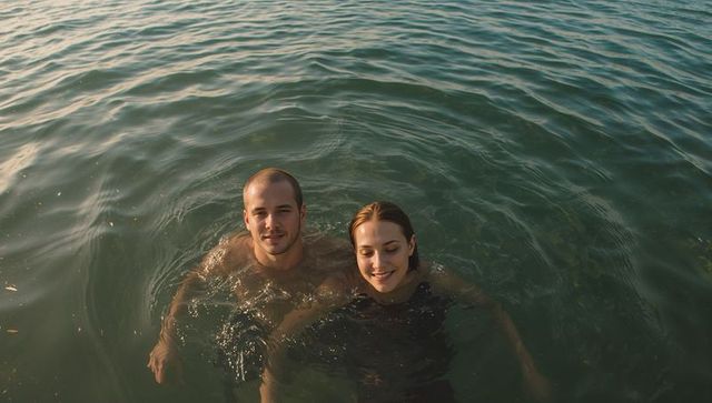 Couple floating in calm open water at golden hour, aerial view of serene summer swim