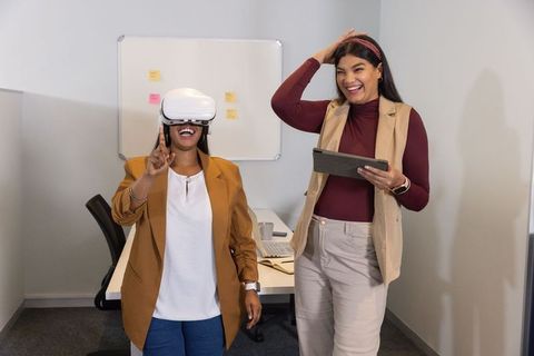 Female coworkers testing vr headset in office, laughing and collaborating
