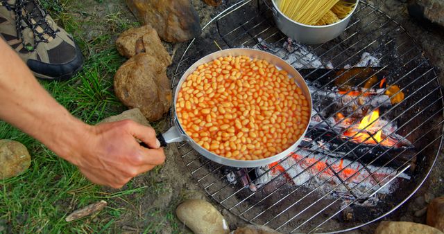 Hand preparing baked beans over a campfire in an outdoor camp setting. Ideal for representing outdoor activities, camping trips, survival skills, nature retreats, bushcraft education, cooking tutorials, and travel adventure promotions.
