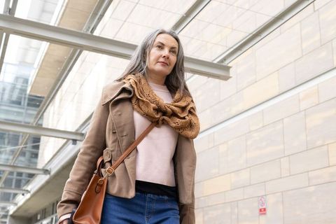 Walking mature woman in brown jacket and tan scarf carrying leather bag under glass canopy