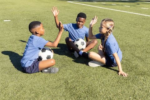Diverse children sitting on soccer field high-fiving while holding soccer balls