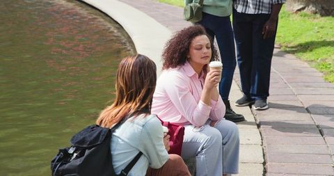 Multiracial friends sitting by curved park pond holding coffee cups, relaxing outdoors