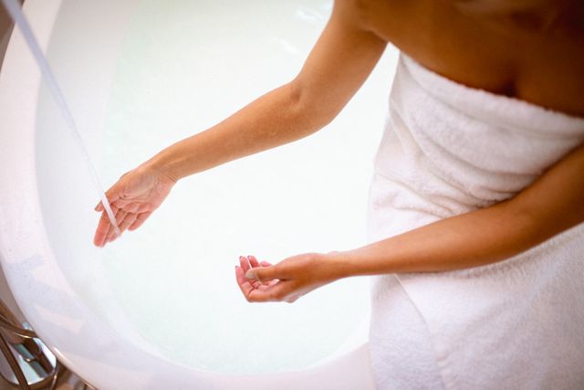 Midsection of biracial woman wearing a robe, sitting on the edge of a bathtub, pouring water for a bath. Ideal for concepts related to self care, relaxation, domestic lifestyle, and wellness. Suitable for use in articles, blogs, and advertisements promoting home spa experiences, hygiene products, and leisure activities.