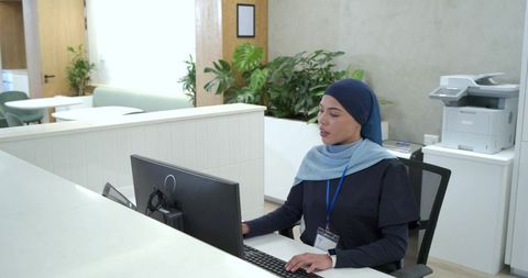 African american receptionist wearing hijab typing at modern clinic reception desk with id badge