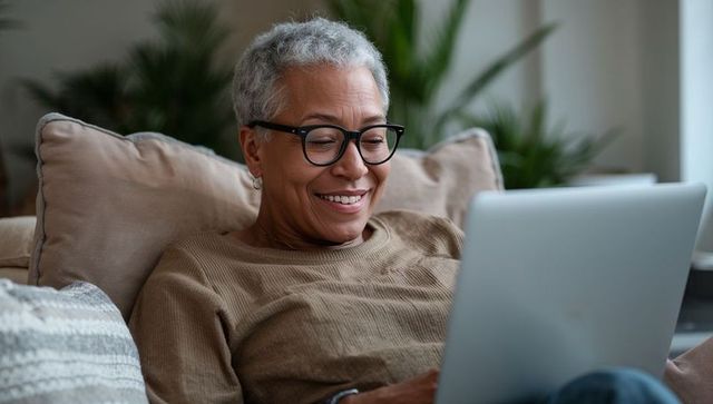Smiling senior woman using laptop on sofa in cozy sunlit living room with houseplants