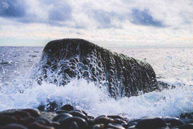 Waves crashing against a steep rock on a shoreline under a cloudy sky depict nature's raw power. Useful for nature-themed projects, coastal travel brochures, or backgrounds emphasizing dramatic seascapes. Perfect for environmental discussions and promoting coastal activities.