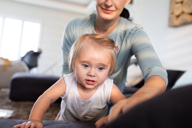 Mother watching baby daughter climbing on couch in living room. Ideal for use in parenting blogs, family lifestyle articles, and advertisements for home safety products. Highlights themes of family bonding, child development, and home life during quarantine.