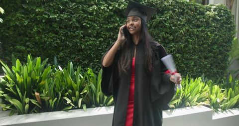 Indian woman graduate talking on phone holding diploma wearing gown and mortarboard in courtyard