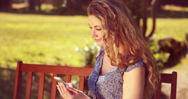 Young woman with long curly hair texting on mobile phone while sitting on a park bench on a sunny day. Ideal for use in promotional materials related to outdoor activities, technology usage, summer lifestyle, or social media apps.