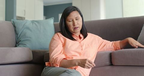 Mature asian woman relaxing on gray sofa in cozy minimalist living room with teal cushion
