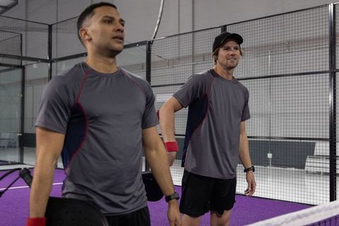 Padel doubles partners standing at net on purple court holding black paddles