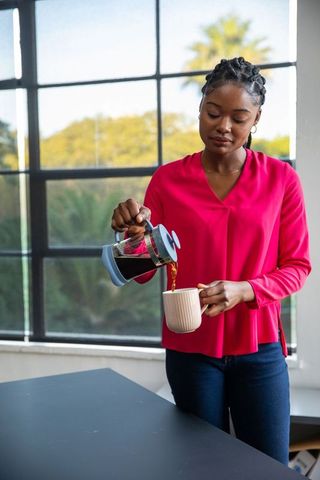 Woman pouring coffee from french press at home by bright window