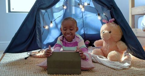 Smiling african american girl using tablet in cozy navy play tent with string lights