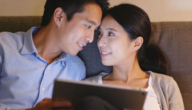 Asian couple cuddling on sofa using tablet in cozy modern living room intimate moment