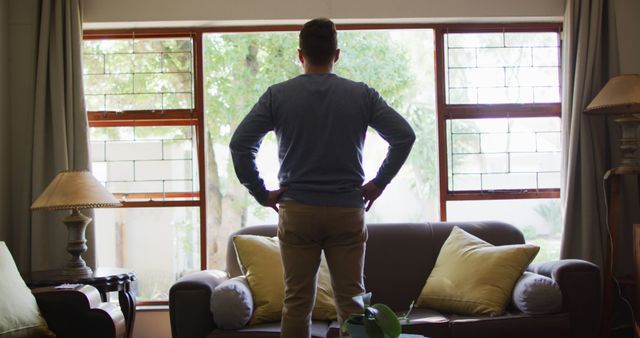 Man is seen standing in front of a living room window with hands on hips, looking outside during the day. This image can be used to depict solitude, contemplation, home environment, or reflection themes.