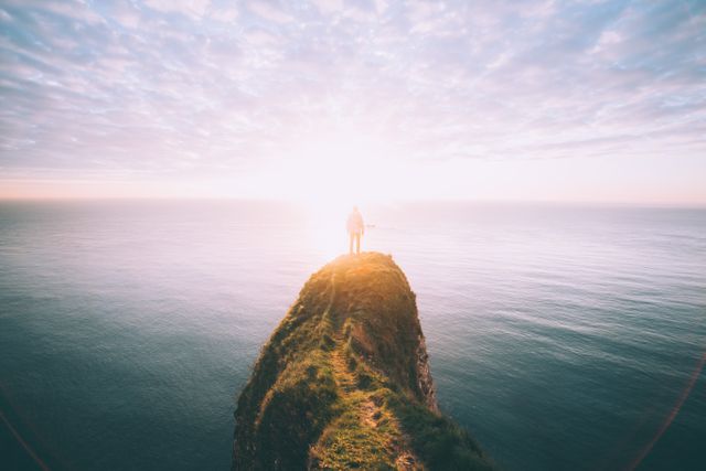 Person standing at the edge of a cliff overlooking vast ocean at sunset. Radiant sunlight creates peaceful and serene atmosphere perfect for use in travel-themed content, motivational posters, inspirational messages, and nature observation materials.