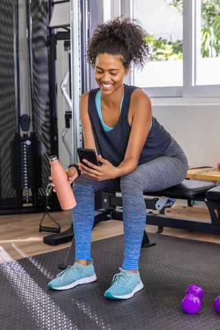 Young woman in gym checking smartphone sitting with water bottle