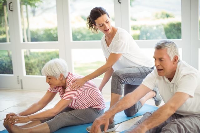 Female trainer assisting senior couple with stretching exercises at home. The elderly man and woman are on yoga mats, focusing on flexibility and fitness. Ideal for use in articles or advertisements about senior fitness, personal training, healthy aging, and home workout routines.