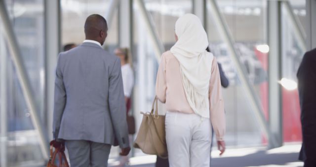 A man in a suit and a woman wearing a hijab and carrying a bag walk together through a modern office corridor. Both individuals dressed in business attire suggest a professional setting and partnership. Ideal for depicting concepts of modern workplace diversity, teamwork, and corporate life in promotional materials, websites, or brochures.