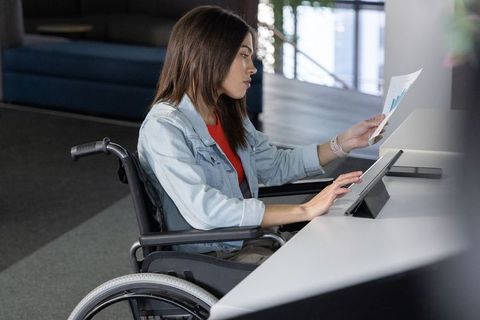 Young woman in wheelchair analyzing charts and using tablet at modern coworking desk